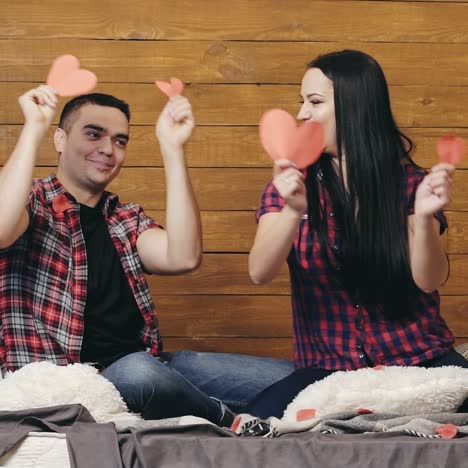 funny couple are sitting on the bed and playing with red paper hearts in their hands on valentine's day