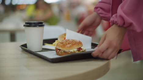 lower body view of lady in pink dress carrying tray with burger, fries, and drink walks towards her table, places it down, and adjusts it before sitting, other people are in the restaurant