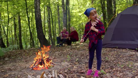 Child-girl-eating-cooked-fried-sausages-near-campfire,-senior-grandparents-on-background-in-wood