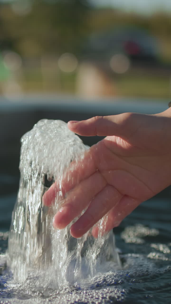 de perto de um homem estendendo a mão para tocar a água de nascente que flui de uma pequena fonte, com reflexos de luz solar na superfície da água e cenário ao ar livre desfocado, incluindo vegetação e banco de parque distante