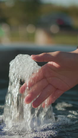 de cerca de un hombre extendiendo la mano para tocar el agua de manantial que fluye de una pequeña fuente, con reflejos de luz solar en la superficie del agua y un entorno al aire libre borroso que incluye vegetación y un banco de parque lejano