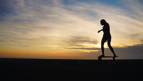 In-a-slow-motion-depiction,-a-woman-skateboards-along-a-road-at-sunset,-framed-by-mountains-and-a-beautiful-sky.-Her-attire-includes-shorts