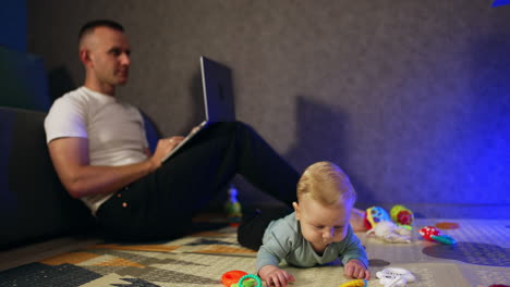 beautiful infant with fair hair lies on the floor. father sits behind leaning on the sofa and working on laptop.