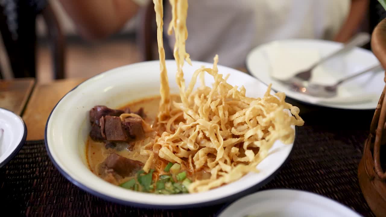 A person uses chopsticks to lift crispy noodles from a bowl of beef curry noodle soup, shot in natural light at a Bangkok eatery