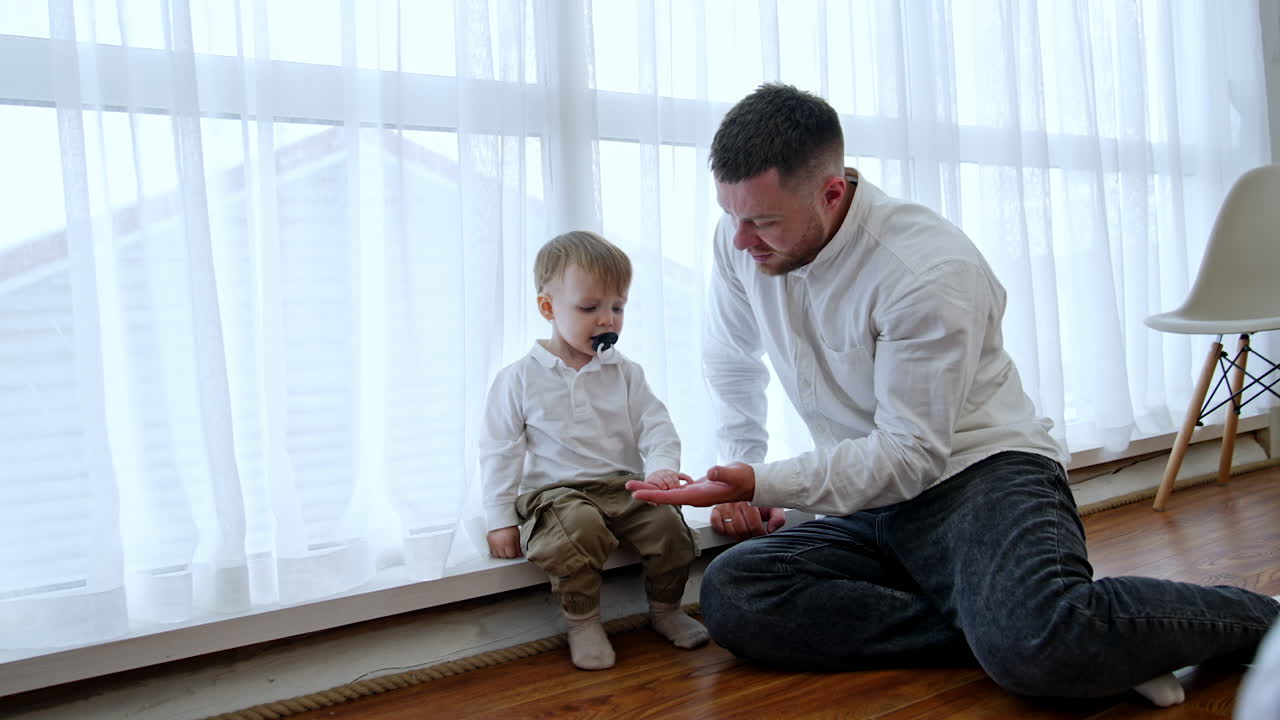 Dad and his little baby boy sit at the window. Toddler with pacifier in mouth stands up and walks to bed.