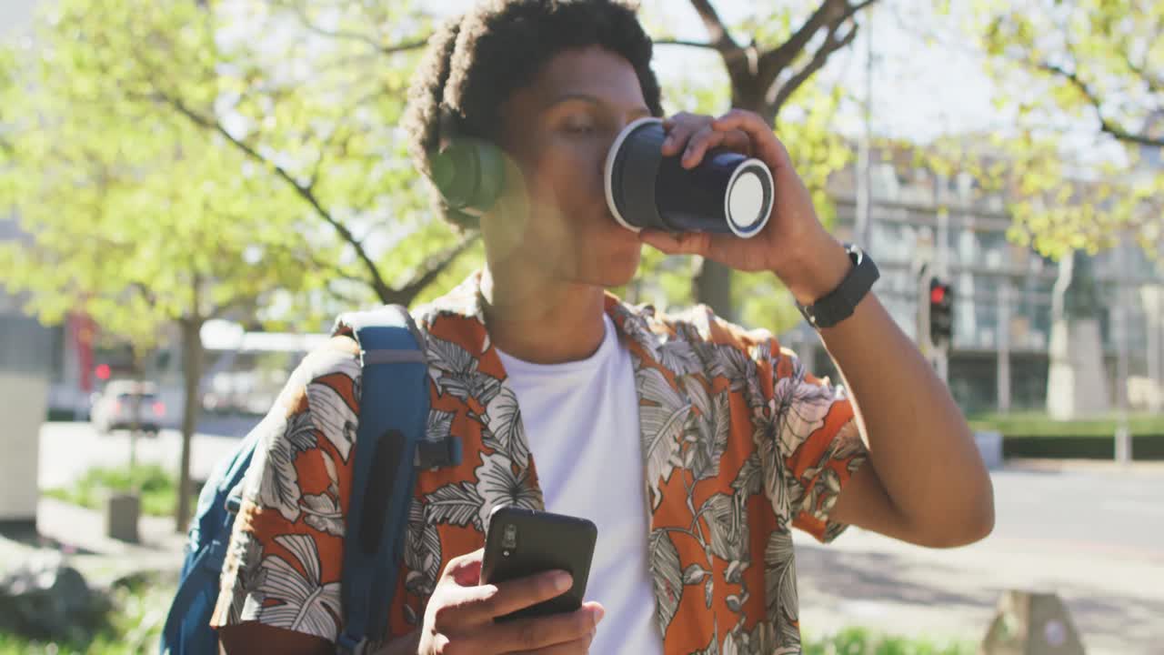 feliz hombre afroamericano en la ciudad, usando teléfono inteligente y auriculares, bebiendo café en la calle