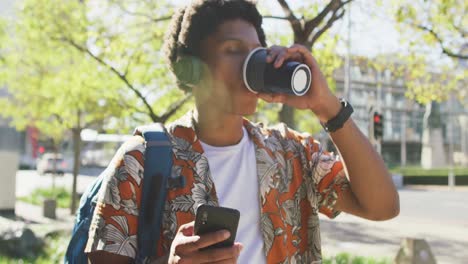 happy african american man in city, using smartphone and headphones, drinking coffee in street