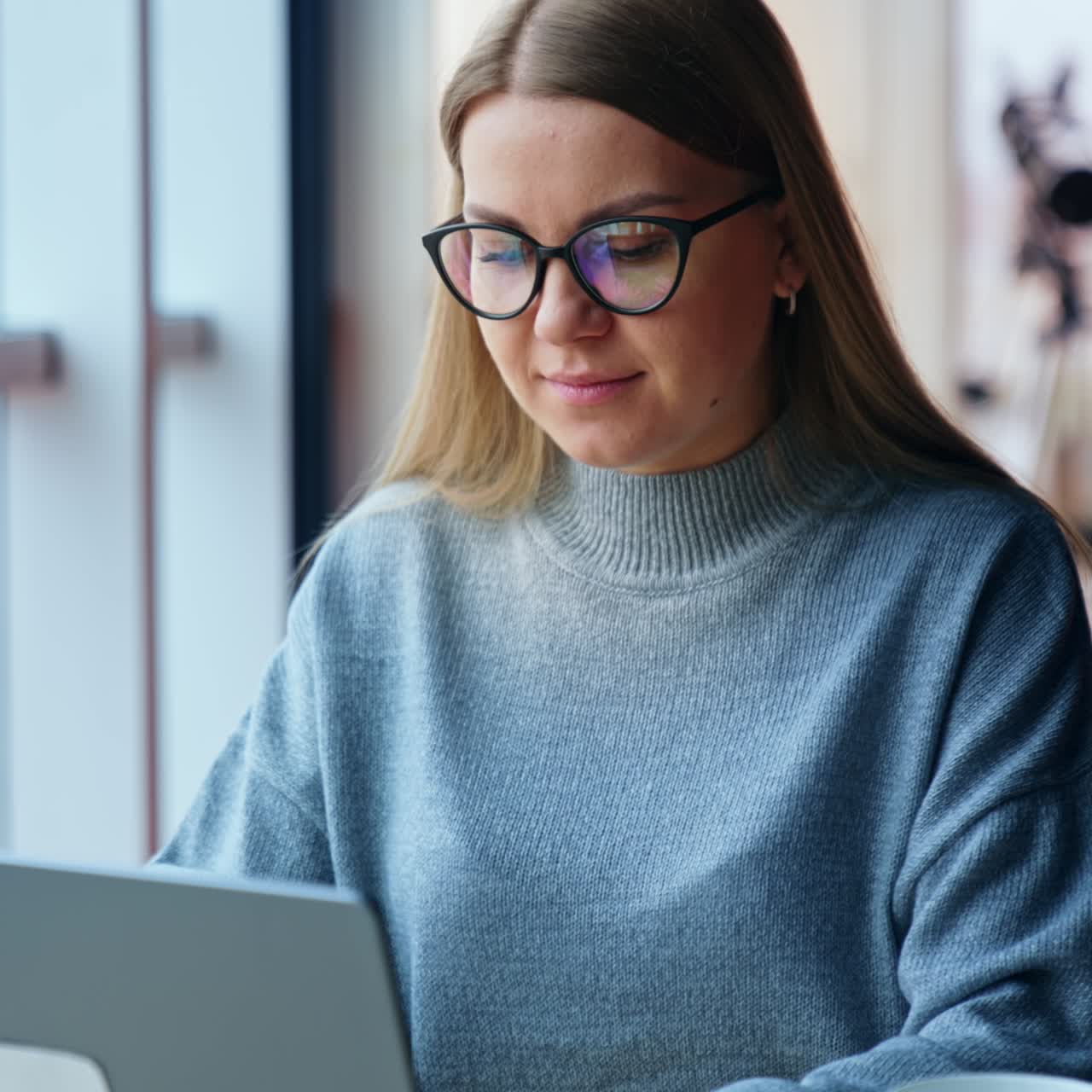 Blonde lady wearing glasses is focused on her work on laptop. Woman is typing something, then looks at camera and smiles. Blurred backdrop