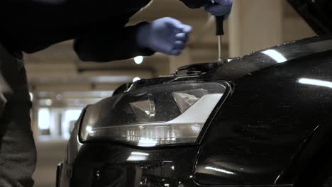 a person unscrewing the screws of a headlight glass cover from a car at a parking lot or garage, and removing the broken light, all while wearing protective gloves