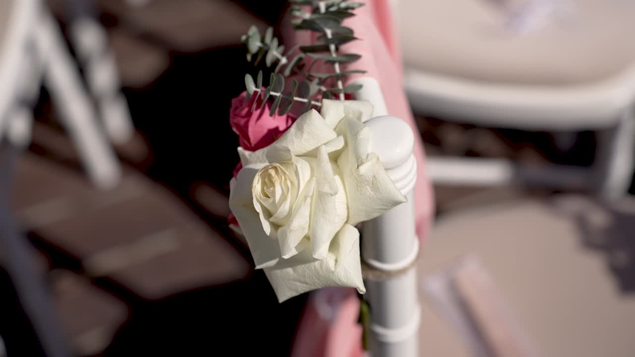 Fresh flowers are attached to white chairs in a garden. The roses add color and charm to the outdoor setup for a special gathering taking place in the daytime