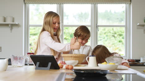 children in pyjamas making pancakes in kitchen at home following recipe on digital tablet