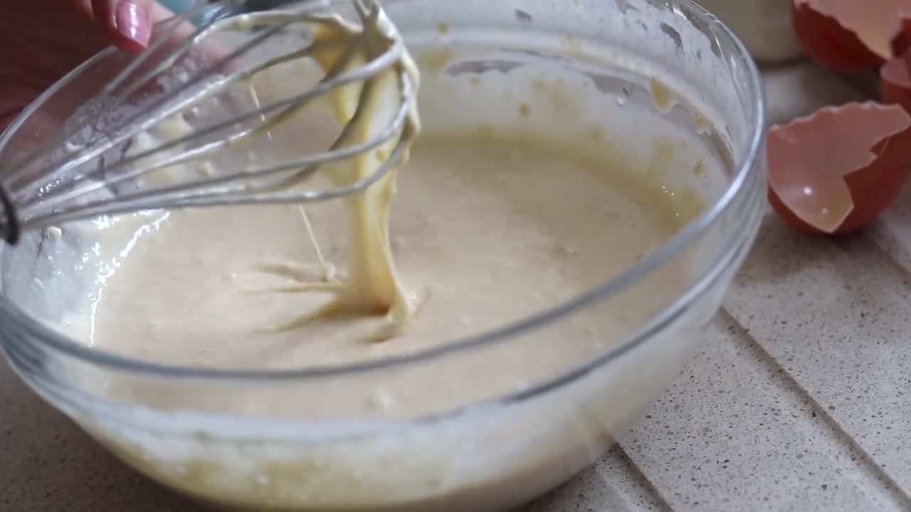 Primer plano de batir masa a mano, en un tazón de vidrio sobre una encimera de cocina, con cáscaras de huevo rotas al fondo