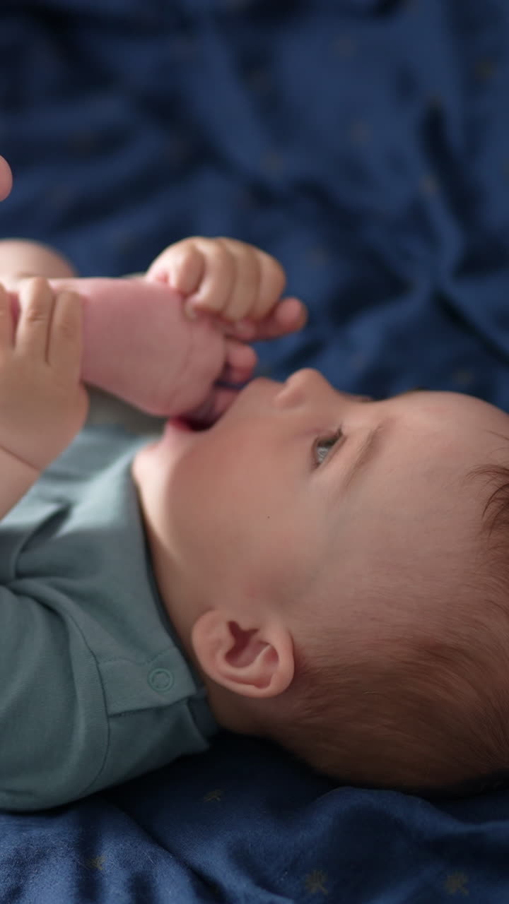 Lovely barefoot child lies on the blue bedclothes. Cute boy pulls his foot to the mouth and chews his toes. Close up. Vertical video