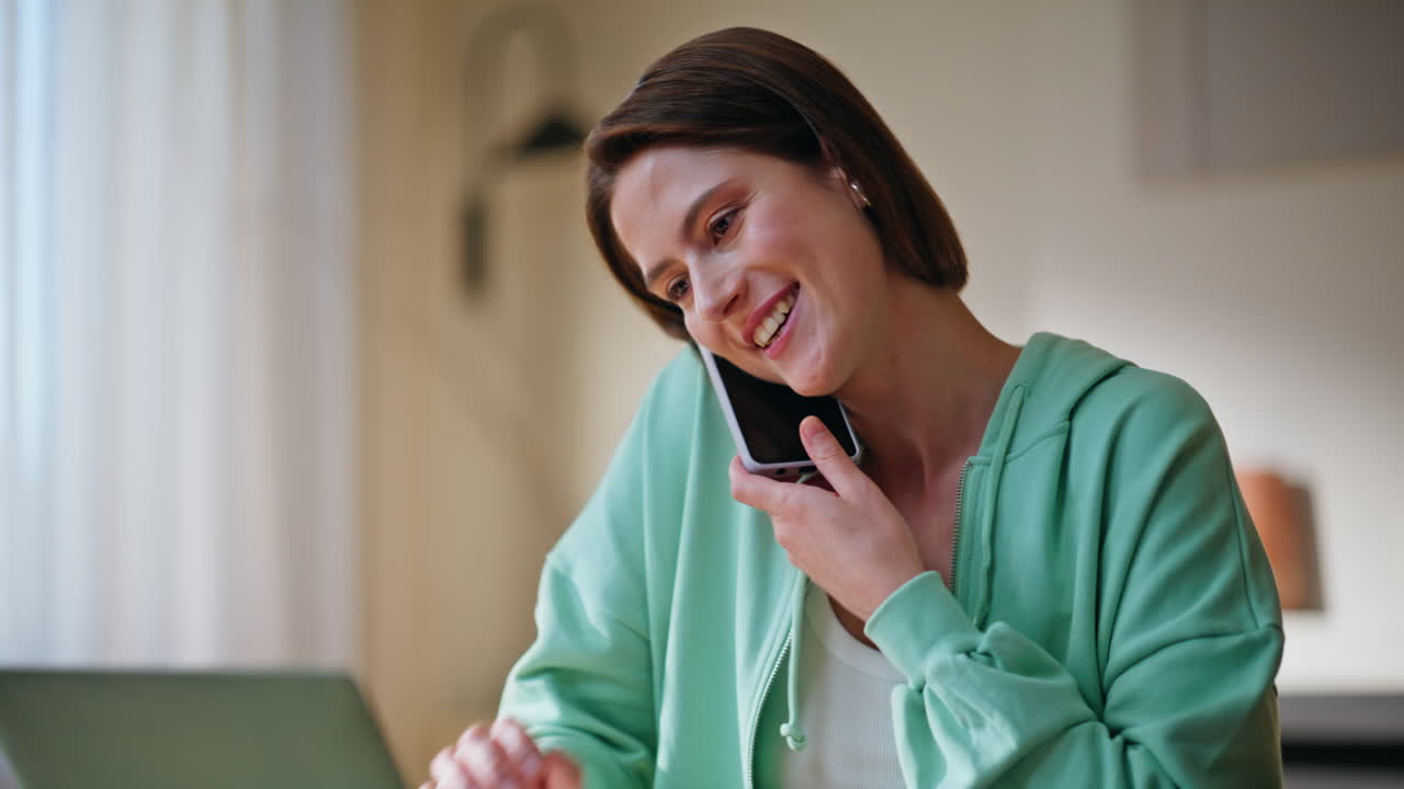 Smiling girl talking mobile phone typing laptop at modern apartment closeup