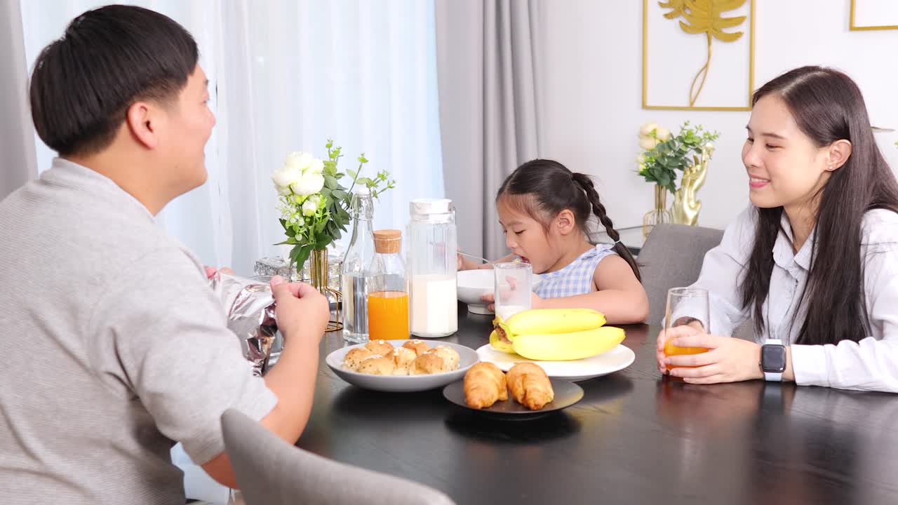 Parents and child share morning meal with milk, juice, bananas, pastries in sunlit home kitchen