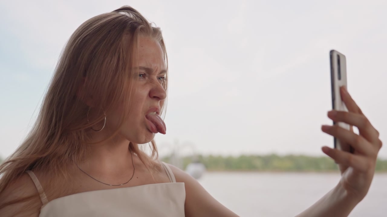Young woman takes selfies outdoors near body of water holding her smartphone and making playful expressions. Woman wears light colored top with thin straps against open sky backdrop