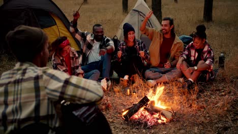 Un-Hombre-Con-Sombrero-Toca-La-Guitarra-Y-El-Resto-Del-Grupo-De-Excursionistas-Toca-Con-él-Y-Se-Divierte-Y-Canta-Mientras-Está-Sentado-Cerca-De-Un-Pequeño-Fuego-En-El-Bosque.-Un-Grupo-Alegre-Y-Feliz-De-Participantes-De-Una-Caminata-Se-Sienta-Cerca-Del-Fuego-Y-Se-Divierte-Con-Canciones-Y-Guitarra.