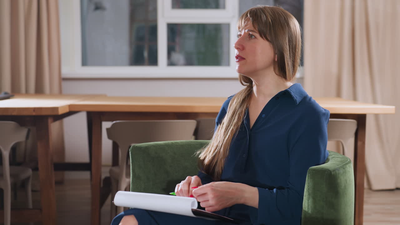 Wellness guide seated in green chair speaking intentionally to guest while holding pen and notebook on lap, warm modern therapy room with wooden table and muted curtains visible in background