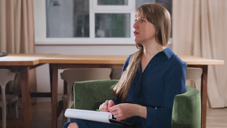 wellness guide seated in green chair speaking intentionally to guest while holding pen and notebook on lap, warm modern therapy room with wooden table and muted curtains visible in background