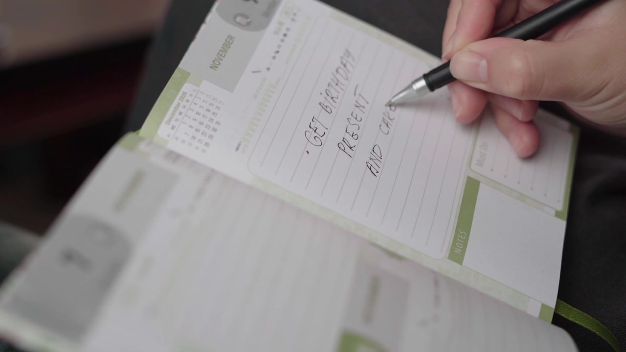 Donna scrive un promemoria per comprare un regalo di compleanno e una torta in un'agenda settimanale personale, focalizzata sulla pianificazione di regali e feste.