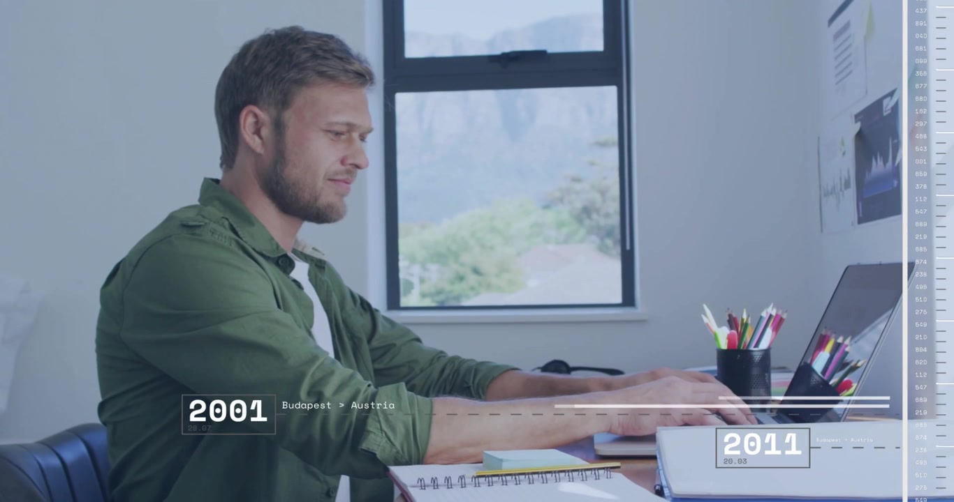 Homem de camisa verde digitando, inclinado em direção ao laptop em uma mesa de escritório, com linha do tempo, caderno e porta-canetas.