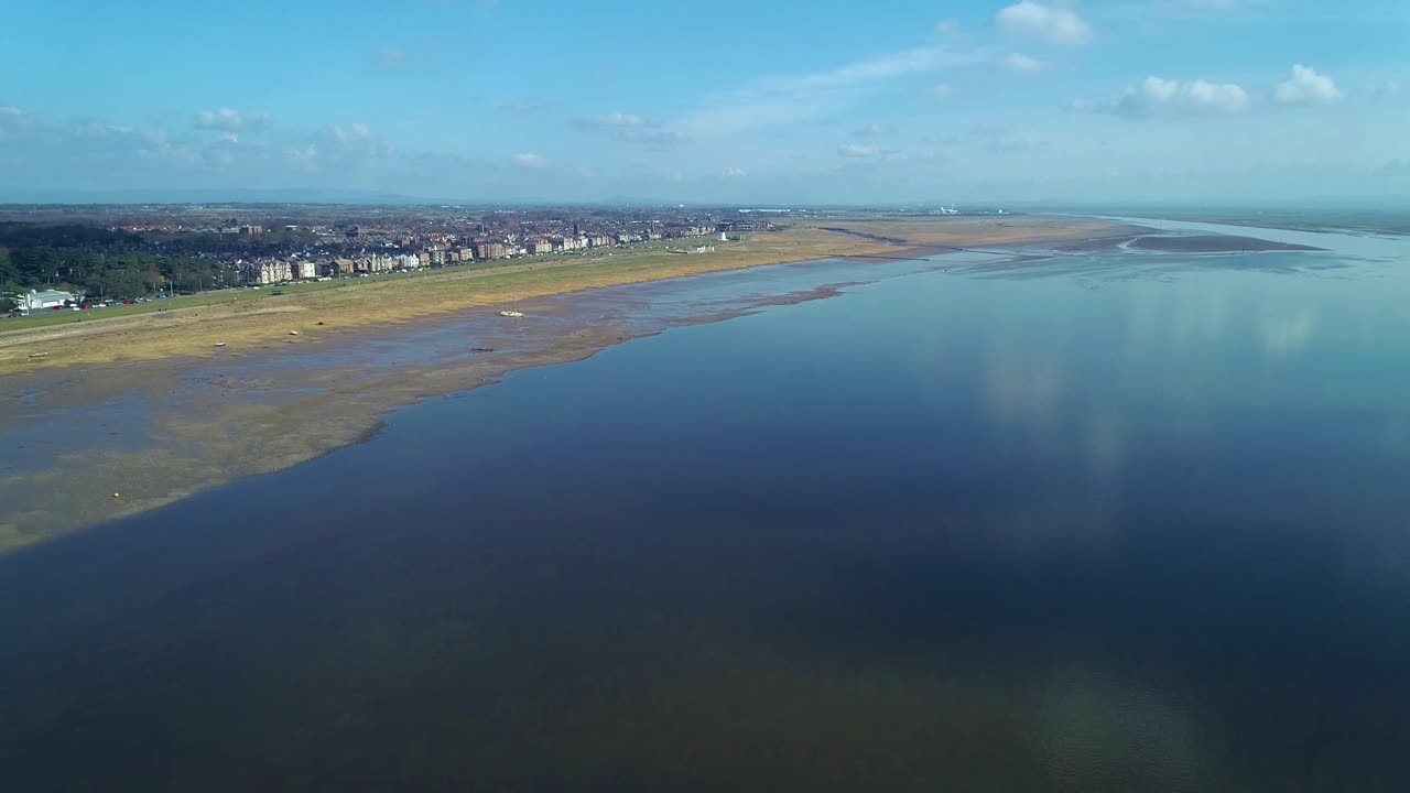 Drone flight towards Lytham shore. Slow dolly towards coastal town on shoreline. The tranquil water is reflective and looks blue due to the blue sky. Lytham is a popular seaside town