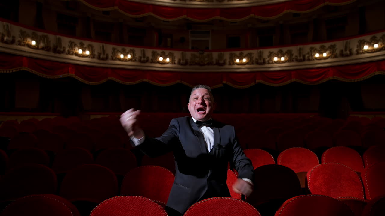 Happy actor in dark theatrical hall. Talented man in suit standing among vacant red chairs and shouting cheerfully. Portrait of a comedian in empty theater.