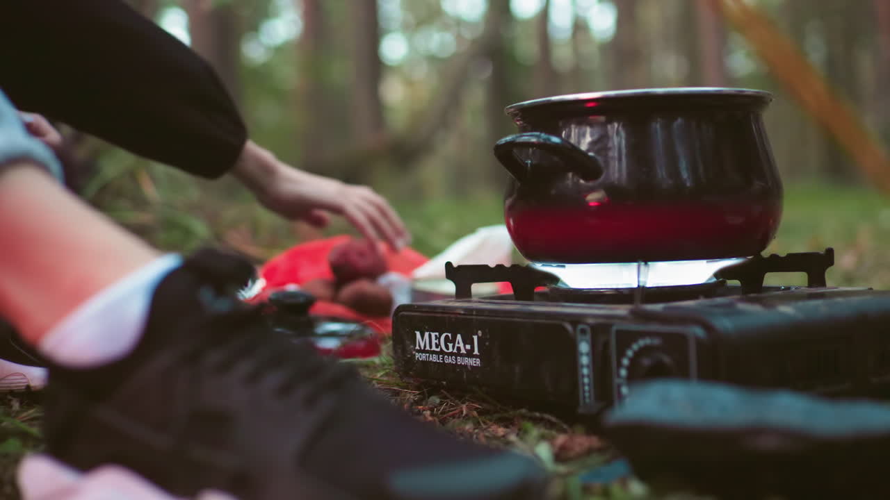 close up of person turning on gas stove under black pot while another pours water into it during outdoor forest cooking session with camping gear visible in natural woodland environment