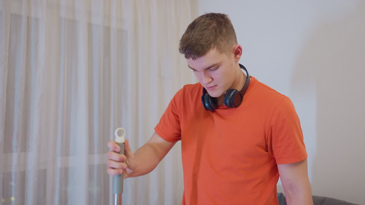 Young man in orange shirt with headphones around neck dipping mop into water bucket and lifting it out during home cleaning routine, demonstrating effort, focus, and sanitation process