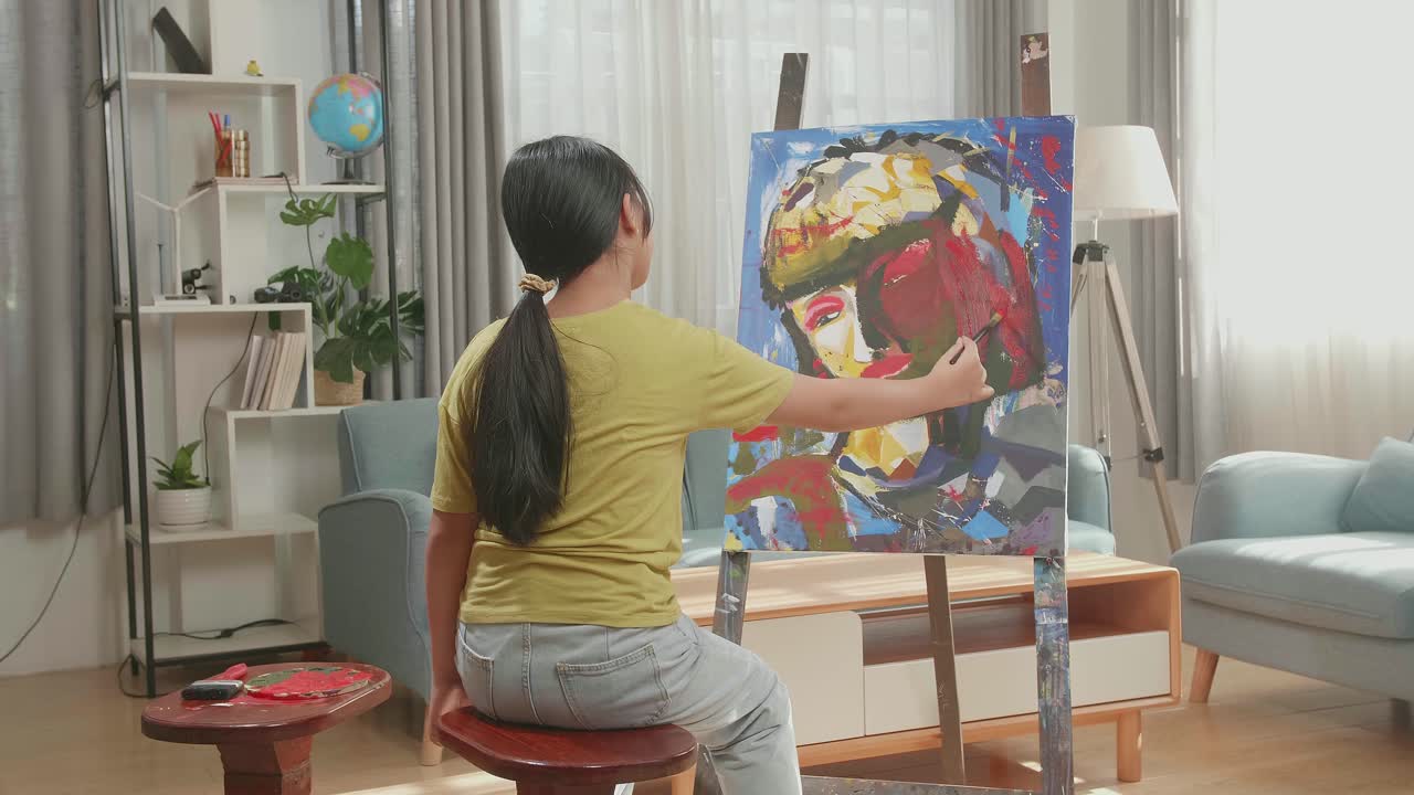Hind View Of An Asian Artist Girl Sitting On Round Wooden Chair Without Backrest Holding Paintbrush Mixed Colour And Painting A Girl'S Face On The Canvas