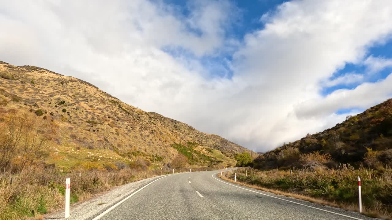 A car travels through Wanaka's winding roads, surrounded by mountainous terrain under a vibrant blue sky with scattered clouds