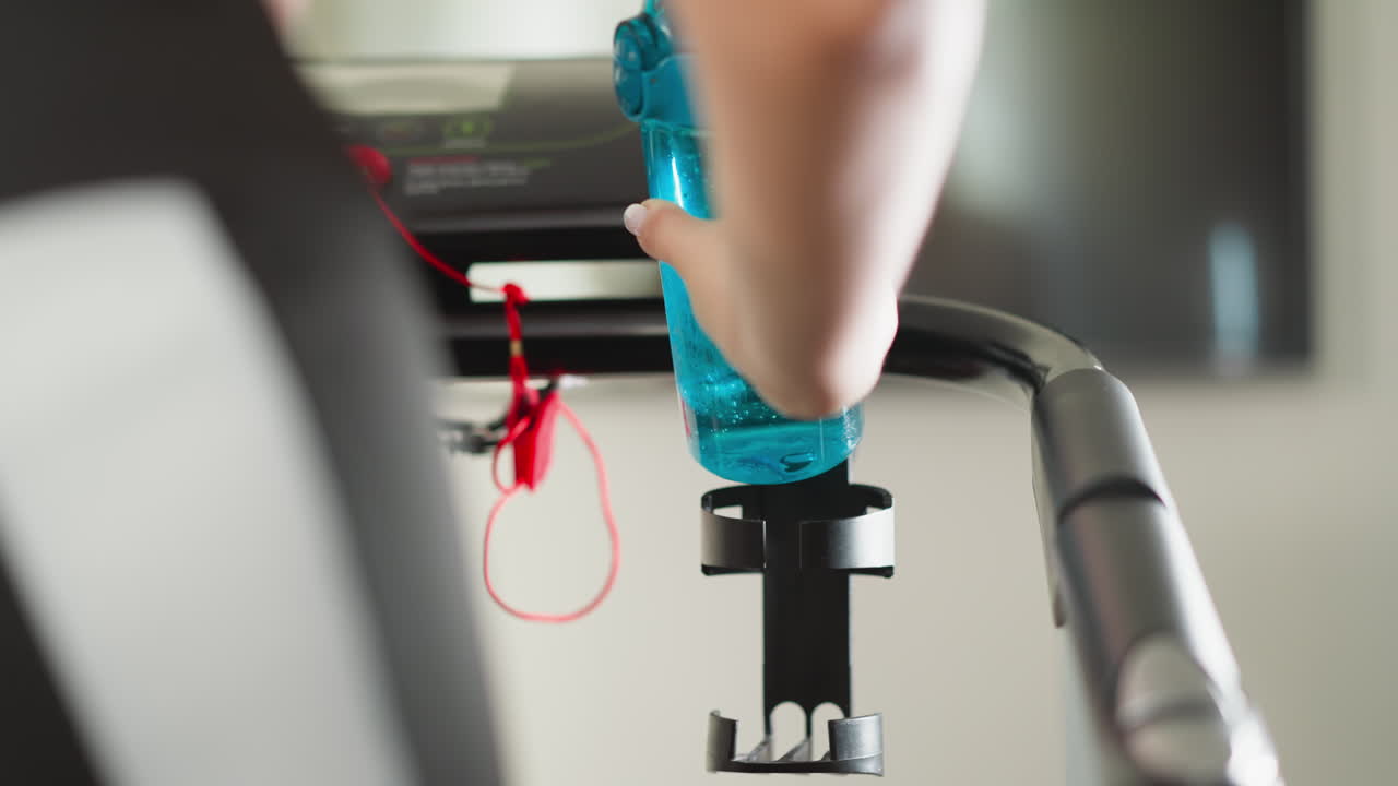 여성은 트레드밀 홀더에 물 병을 다시 놓습니다. 운동선수 여성은 피트니스 클럽에서 물 한 잔을 마신 후 계속 달립니다. 훈련 중에 수분 수준을 유지합니다.