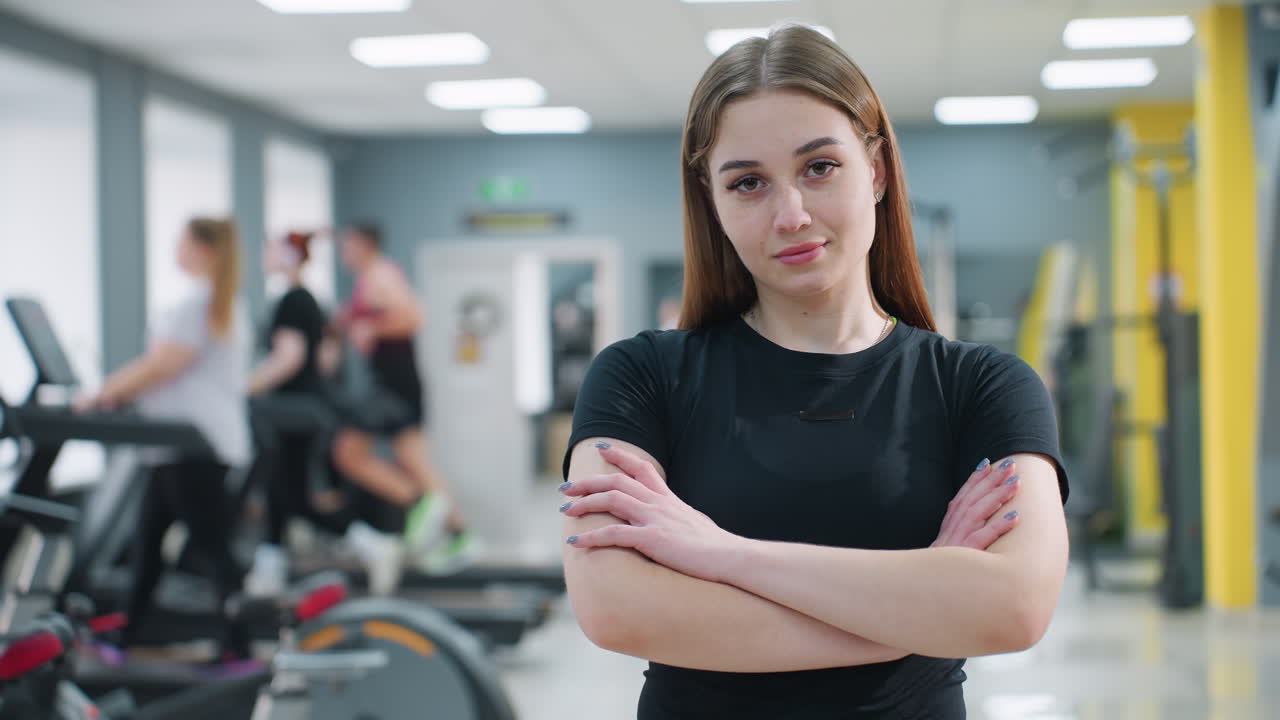 Confident young female athlete posing with arms crossed in fitness center, looking directly at camera with calm expression while people work out on gym equipment in background