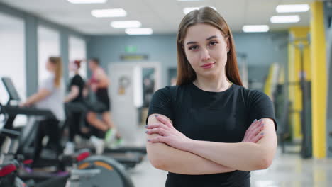 confident young female athlete posing with arms crossed in fitness center, looking directly at camera with calm expression while people work out on gym equipment in background