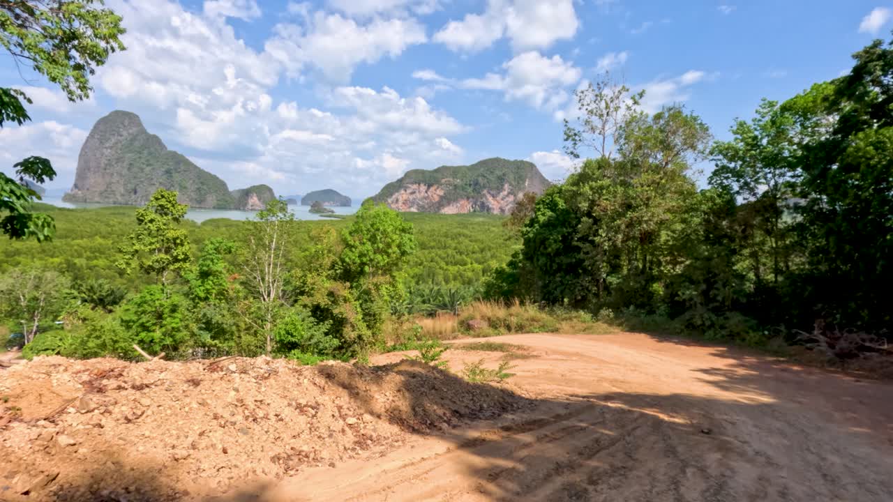 Camera glides along rural dirt road, revealing tropical trees, hills, and dramatic limestone mountains