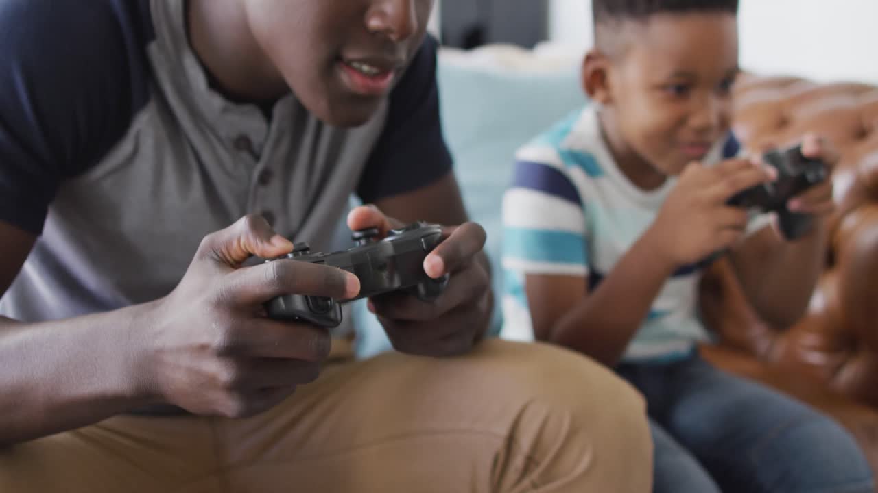Video of happy african american father and son sitting on sofa and playing video games