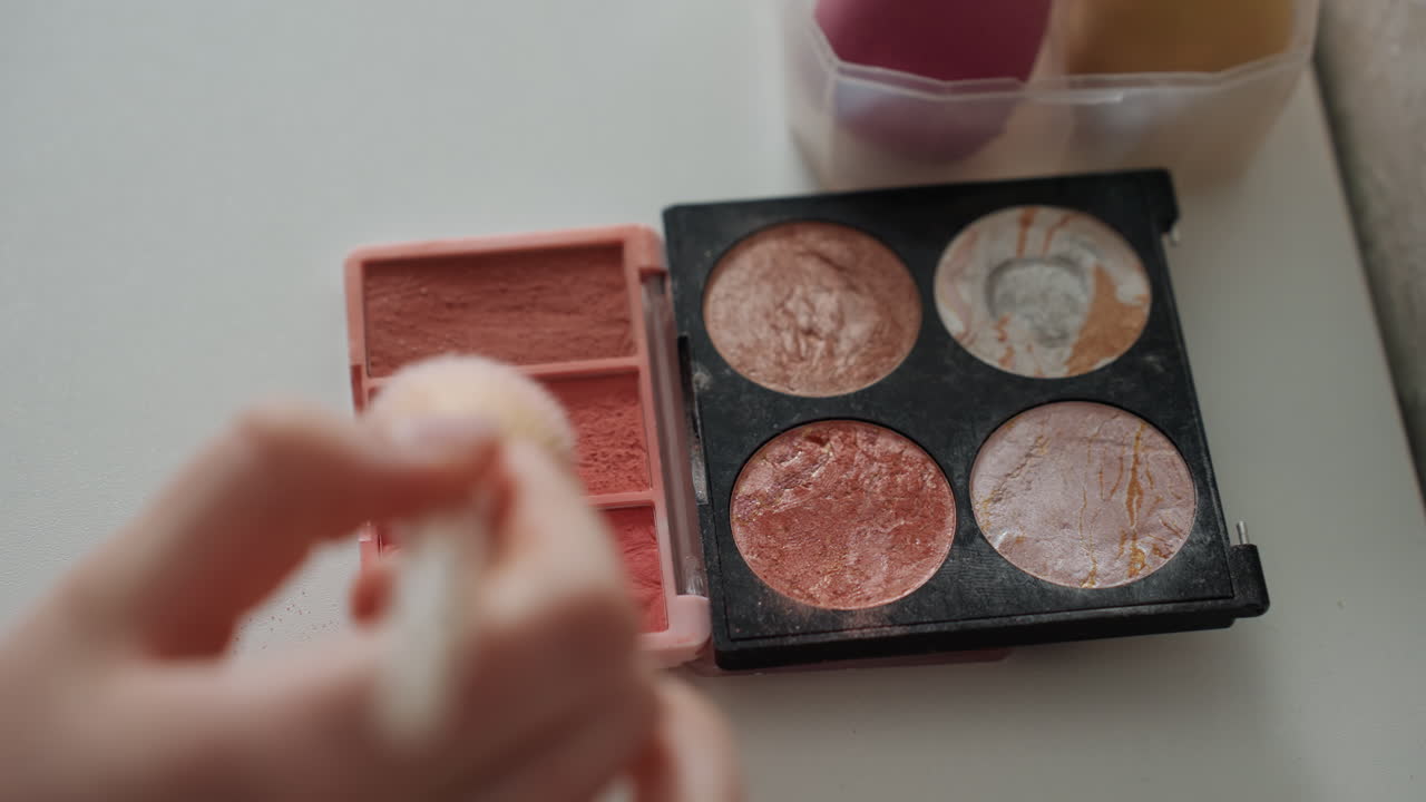 Closeup makeup palette with shimmer pans, hand dabbing sponge into warm copper and peach shades, soft powder fallout on white table, compact with four glitter and matte pans, cup of brushes blurred