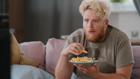 hombre obeso comiendo papas fritas con ketchup mientras ve la televisión en casa