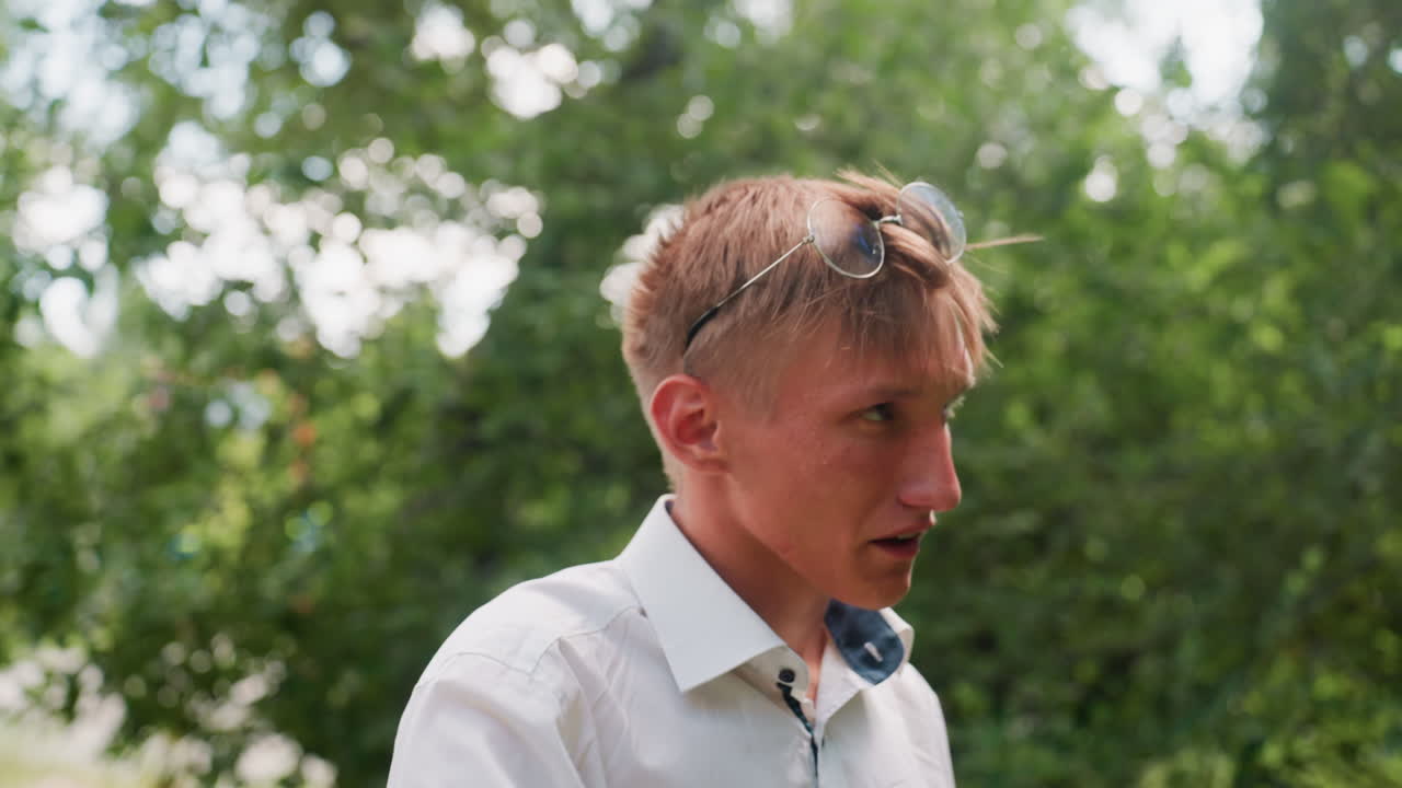 Close up view of young man in white shirt with glasses walking tired and scratching eyes against blurred green forest background, expressing fatigue and discomfort in natural outdoor environment