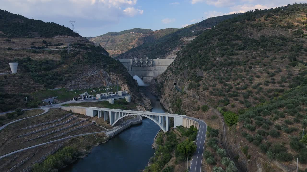 vista aérea de un puente y una represa de río con agua que fluye rodeada de montañas y valles en un día soleado