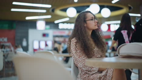 lady with curly hair seated in restaurant, gazing thoughtfully into distance with a calm expression, as people pass by in background, with soft, ambient lighting