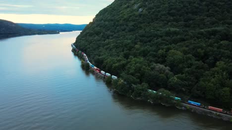 Aerial-drone-footage-of-a-long-freight-train-going-along-the-tracks-at-the-base-of-a-mountain-next-to-a-river-in-the-Appalachian-mountains-at-sunset