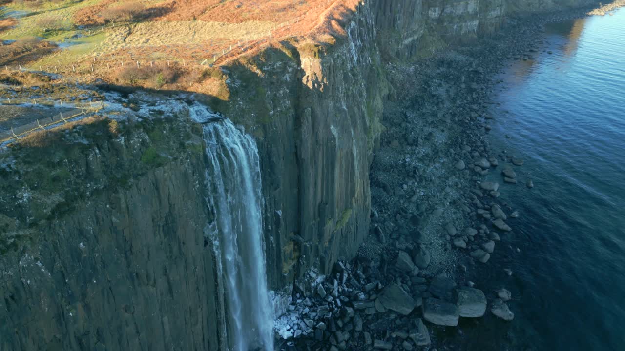 cascada en la sombra en cascada por el acantilado empinado a la playa helada y pedregosa, temprano en la mañana en invierno con la cima del páramo iluminado por el sol en la cascada de kilt rock, isla de skye, highlands occidental, escocia, reino unido