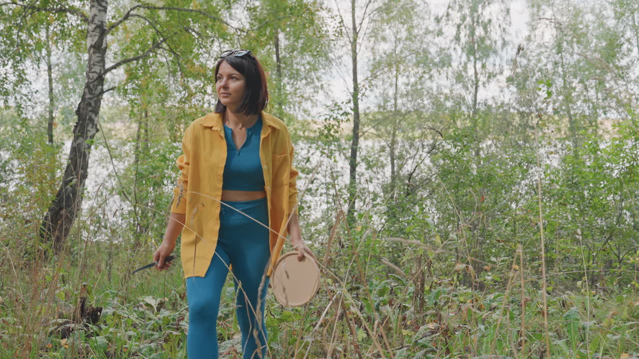 Donna che cammina in una foresta di betulle durante un campeggio, giacca gialla e completo blu, trasportando piccola attrezzatura mentre si muove attraverso il sottobosco verde vicino alla calma del lago, atmosfera serena con alte betulle, erba.