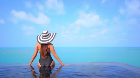 Frau-In-Schwarzem-Badeanzug-Und-Großem-Hut,-Die-Am-Rand-Des-Infinity-Pools-Mit-Blick-Auf-Das-Türkisfarbene-Meer-Sitzt,-Hebt-Die-Arme-Und-Genießt-Den-Blick-Auf-Das-Atemberaubende-Tropische-Resort-Am-Strand