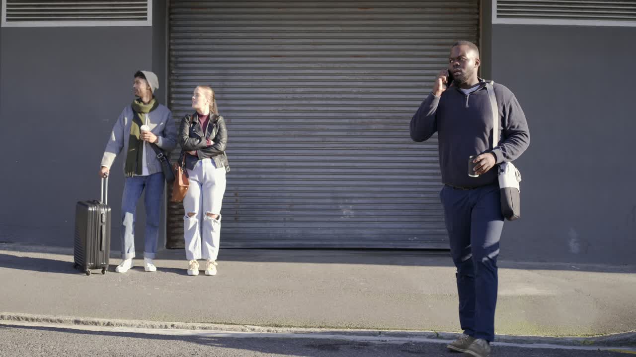 Diverse pair waiting by suitcase at shutter while man following phone call walking and gesturing