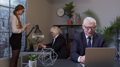 Cheerful-mature-old-businessman-raising-hands-celebrating-sudden-victory-with-colleagues-in-office