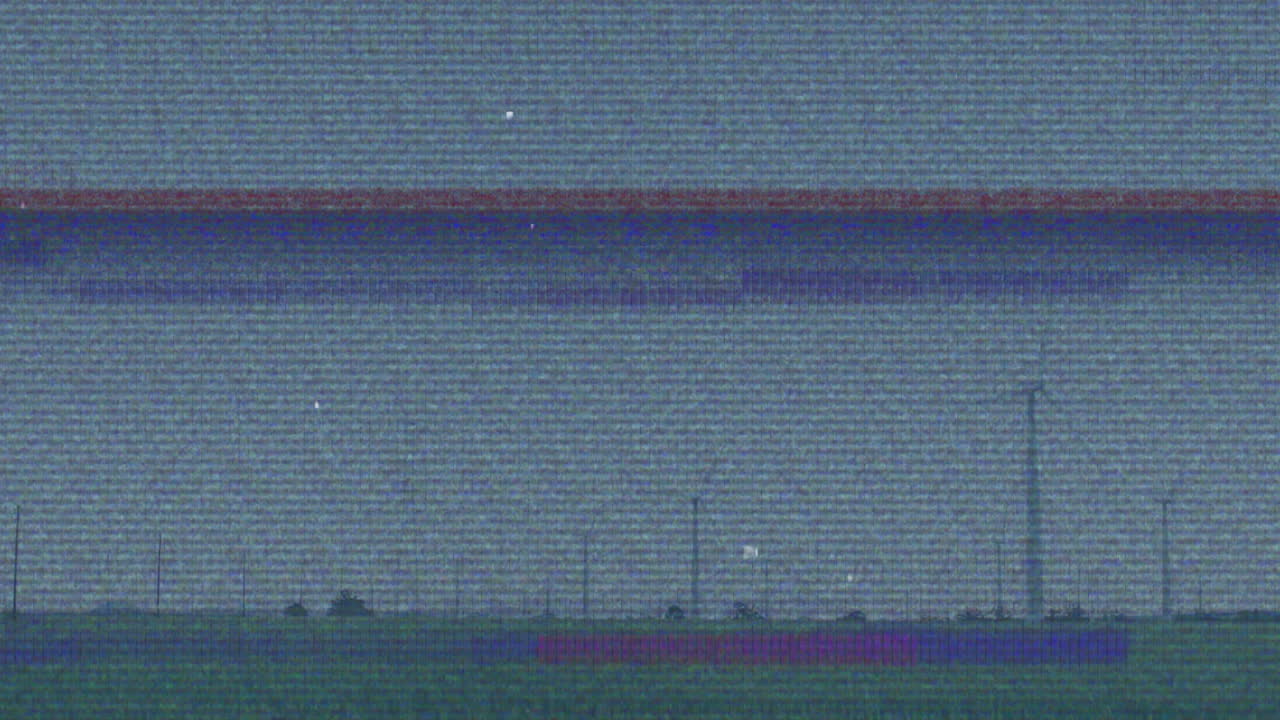 Grainy static panorama showing flat grassy field punctuated by fence posts and turbine silhouette