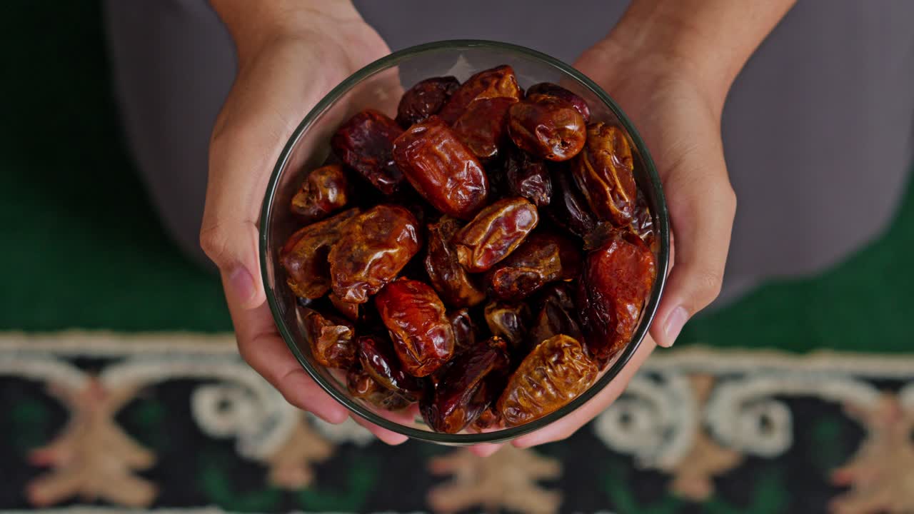Top-down view of hands holding glass bowl of fresh organic dates over prayer mat