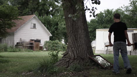 a young high school aged teen boy holding an axe walks up to a mirror laying on the ground